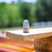Stainless steel 1/4-inch Reaper rotating sewer jetting nozzle standing on a wooden plank outdoors, showing engraved Reaper logo and polished metal finish. High‑pressure drain cleaning nozzle for 2–4 inch pipes with rotating front jet and four rear propulsion jets, built with carbide wear surfaces for durability.