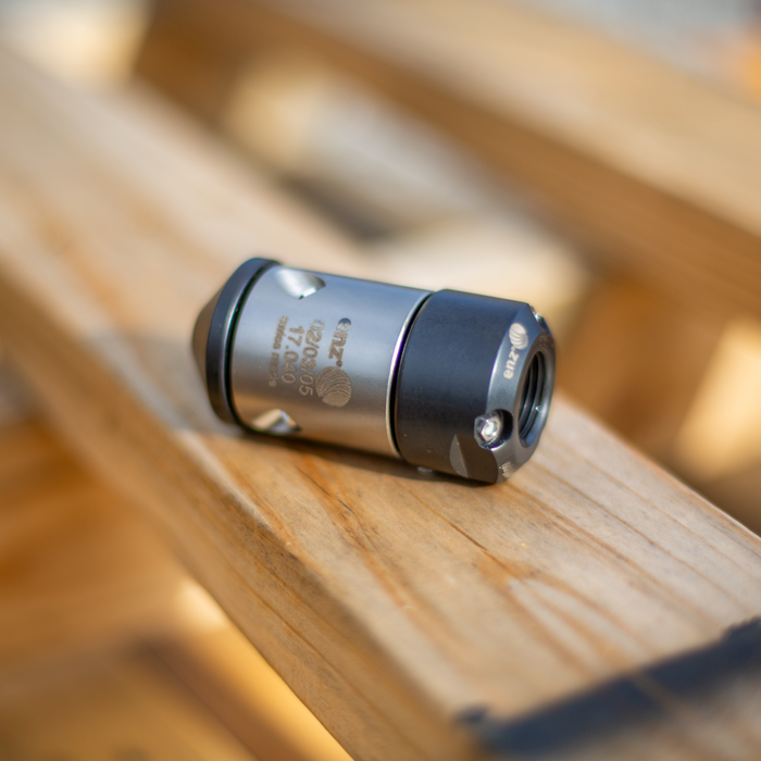 HRV Nozzle - Swiss‑made precision‑engineered metal nozzle with engraved identifiers, featuring a cylindrical body and tapered tip, positioned on a wooden workshop surface; a durable industrial component used in professional mechanical and manufacturing settings.