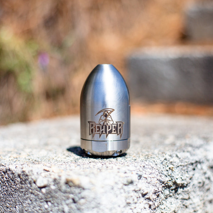 Reaper Rotating Nozzle placed on an outdoor concrete surface, featuring a polished stainless‑steel body with the engraved Reaper logo. This high‑pressure sewer jetting nozzle delivers a powerful forward oscillating jet and four rear propulsion jets to cut blockages, penetrate roots, and clear grease with a cone‑shaped spray pattern. Precision‑machined design ideal for professional drain cleaning, available in multiple pressure and flow sizes from US Jetting.