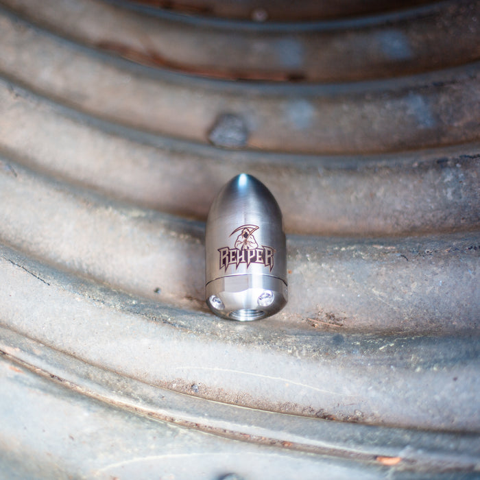 Reaper Rotating Nozzle on a grooved metal surface, featuring a polished stainless‑steel body with the engraved Reaper logo. This high‑pressure sewer jetting nozzle delivers a powerful forward oscillating jet and four rear propulsion jets to cut blockages, penetrate roots, and clear grease with a cone‑shaped spray pattern. Precision‑machined design ideal for professional drain cleaning, available in multiple pressure and flow sizes from US Jetting.