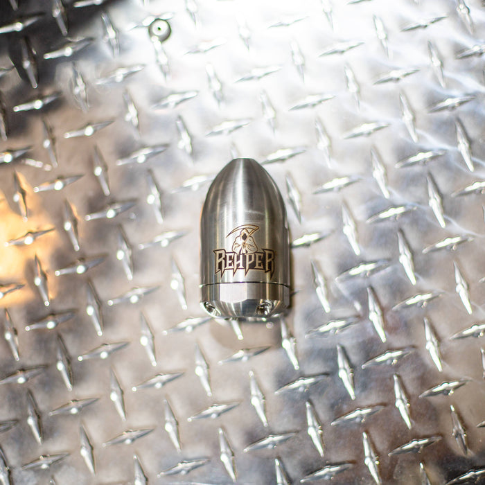 Reaper Rotating Nozzle on a diamond‑plate surface, featuring a polished stainless‑steel body with the engraved Reaper logo. This high‑pressure sewer jetting nozzle delivers a powerful forward oscillating jet and four rear propulsion jets to cut blockages, penetrate roots, and clear grease with a cone‑shaped spray pattern. Precision‑machined design ideal for professional drain cleaning, available in multiple pressure and flow sizes from US Jetting.