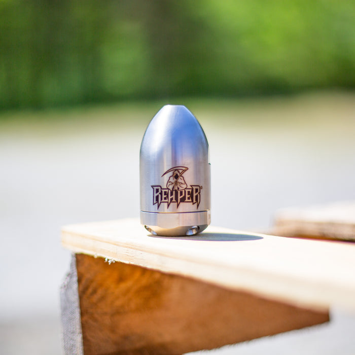 Reaper Rotating Nozzle displayed outdoors on a wooden surface, featuring a polished stainless‑steel body with the engraved Reaper logo. This high‑pressure sewer jetting nozzle delivers a powerful forward oscillating jet and four rear propulsion jets to cut blockages, penetrate roots, and clear grease with a cone‑shaped spray pattern. Precision‑machined design ideal for professional drain cleaning, available in multiple pressure and flow sizes from US Jetting.