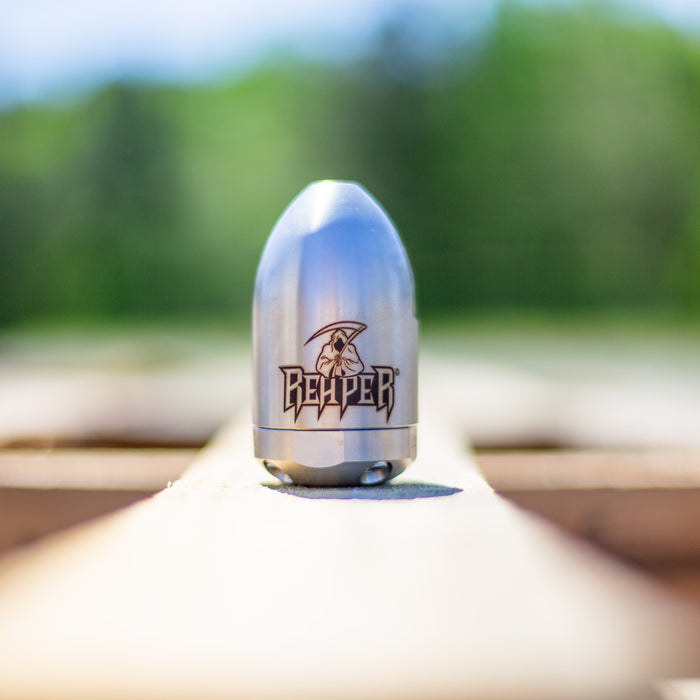 Reaper Rotating Nozzle displayed outdoors on a wooden surface, featuring a polished stainless‑steel body with the engraved Reaper logo. This high‑pressure sewer jetting nozzle delivers a powerful forward oscillating jet and four rear propulsion jets to cut blockages, penetrate roots, and clear grease with a cone‑shaped spray pattern. Precision‑machined design ideal for professional drain cleaning, available in multiple pressure and flow sizes from US Jetting.