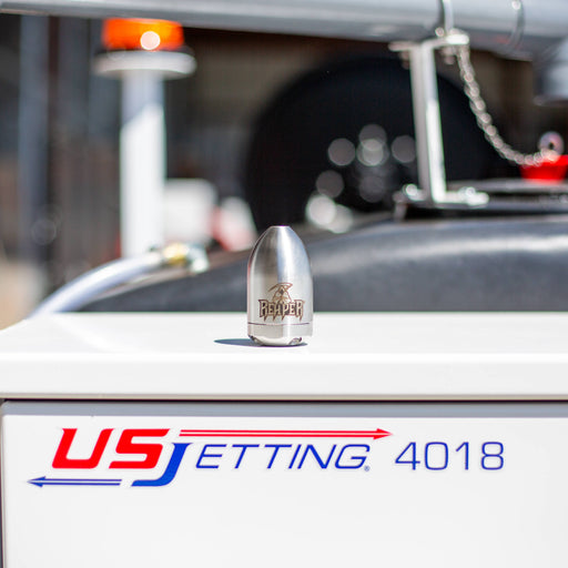 Reaper Rotating Nozzle displayed on a US Jetting 4018 jetter, featuring a stainless‑steel body with the REAPER logo and precision jet ports. This high‑pressure sewer jetting nozzle uses a powerful forward oscillating jet and four rear propulsion jets to cut through blockages, penetrate roots, and clear grease with a cone‑shaped spray pattern. Ideal for professional drain cleaning applications and available in multiple pressure and flow sizes from US Jetting.
